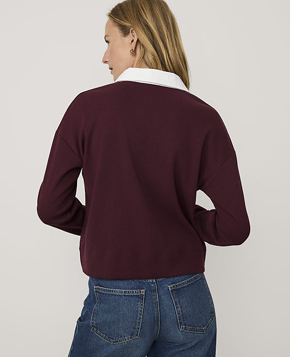 A woman wearing a maroon sweater with a white collar and blue jeans stands against a plain light gray background.