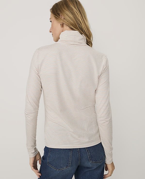 A woman wearing a striped turtleneck and blue jeans stands against a plain white background.