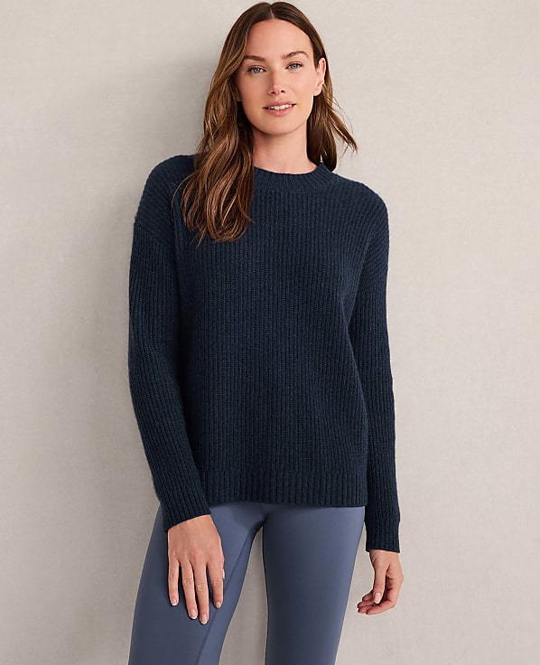 A woman in a light gray ribbed sweater stands against a textured, neutral background.