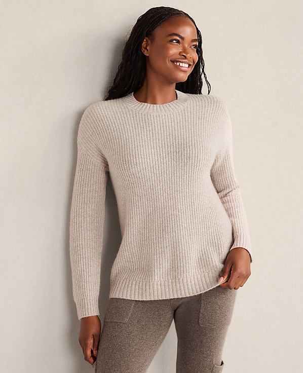 A smiling woman wearing a light beige ribbed sweater and brown pants leans against a plain, neutral-colored wall.