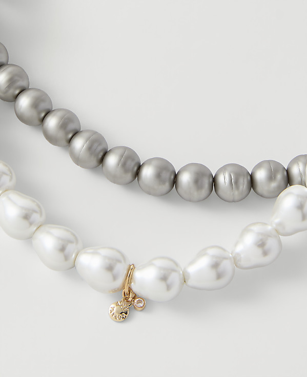 Two strands of pearls, one grey and one white with a small gold charm, rest against a plain light grey background.