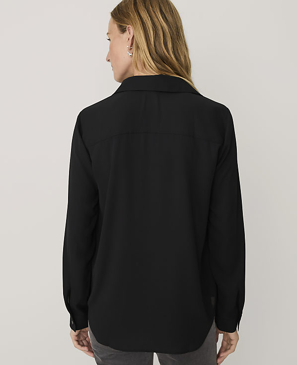A woman wearing a black long-sleeved button-down shirt stands with her back to the camera against a plain light gray background.