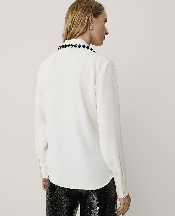 A woman wearing a white long-sleeved blouse with a beaded collar and black sequined pants stands against a plain light gray background.