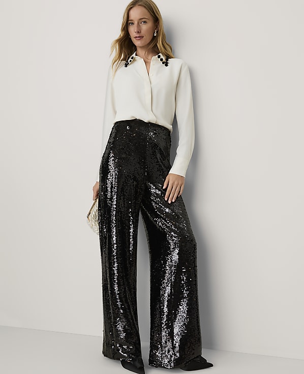 A woman wearing a white blouse and shimmering black sequined wide-leg trousers stands against a plain white wall.