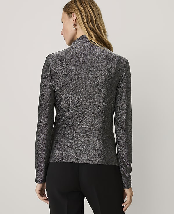 A woman wearing a shimmering grey long-sleeved top and black trousers stands with her back to the camera against a plain white background.
