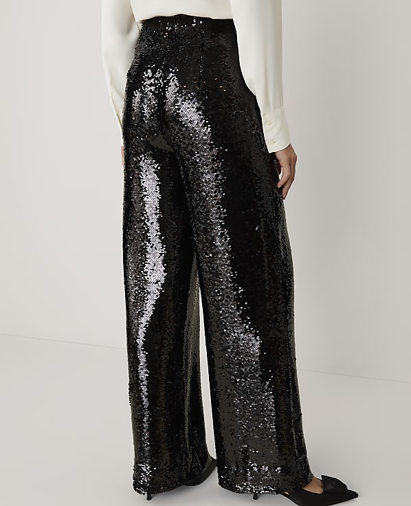 A person wears shimmering black sequined wide-leg trousers and a white blouse against a plain, light-colored background.