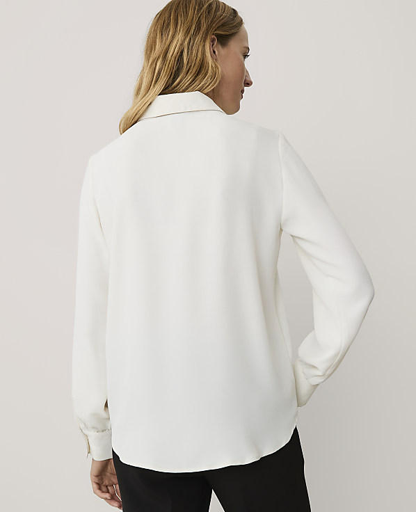 A woman with blonde hair wearing a white long-sleeved shirt and black trousers stands against a plain light grey background.