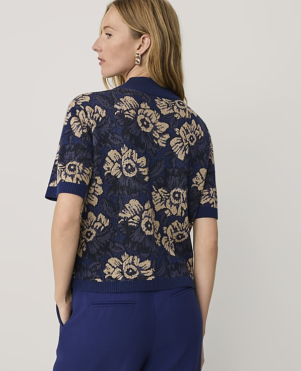 A woman wearing a blue and gold floral knit top and blue trousers stands against a plain light gray background.