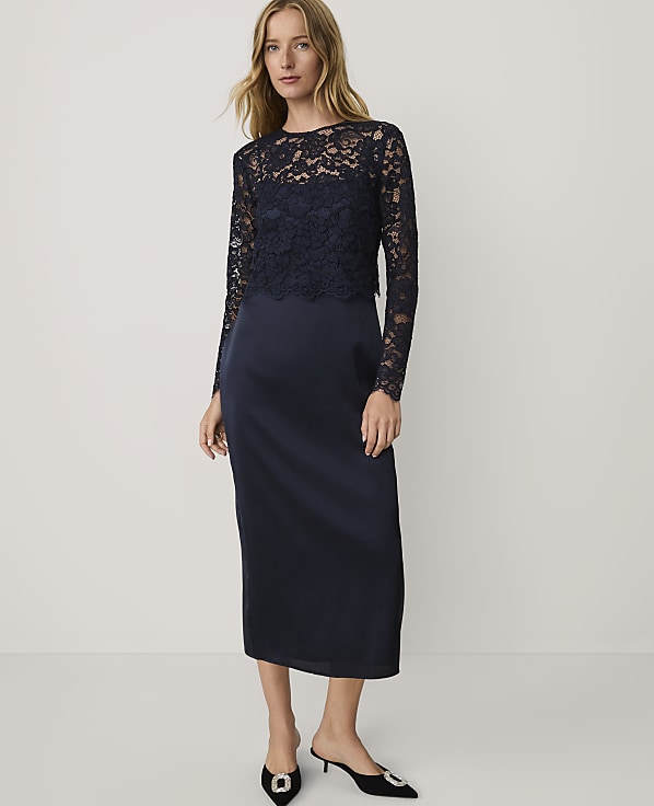 A woman wearing a navy blue lace and satin dress and black heels stands against a plain white background.