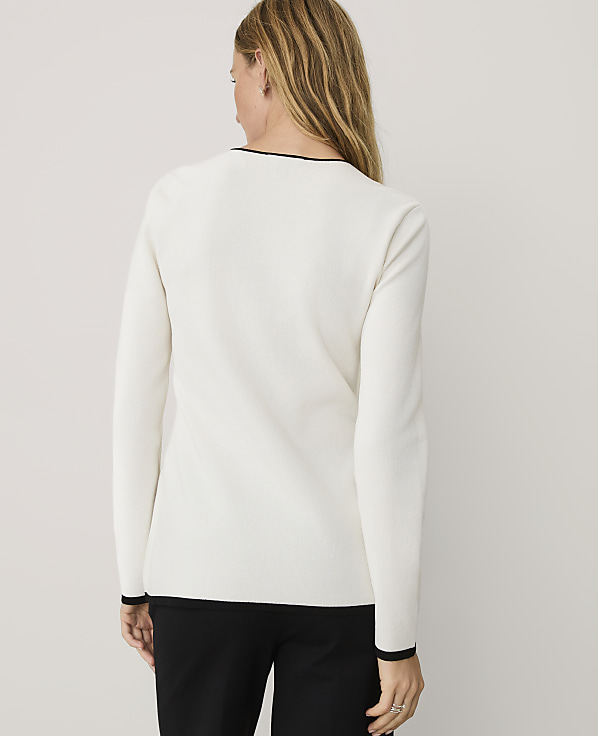 A woman wearing a white long-sleeved top and black trousers stands with her back to the camera against a plain light gray background.