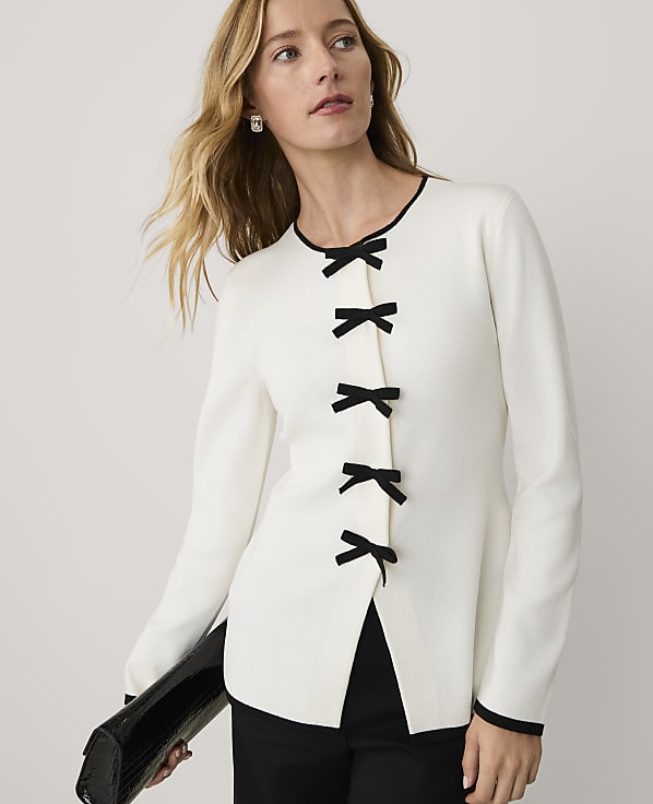 A woman wearing a white long-sleeved top with black bow closures and black trousers stands against a plain white background.