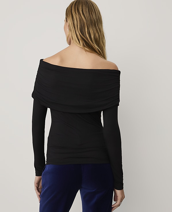 A woman wearing a black off-the-shoulder top and blue velvet pants stands against a plain light gray background.
