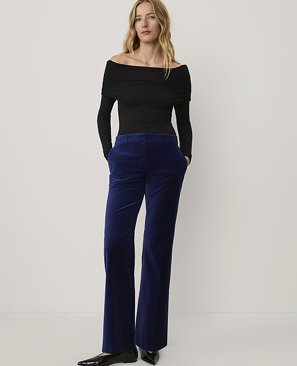 A woman wearing a black off-the-shoulder top and blue velvet trousers stands against a plain white background.