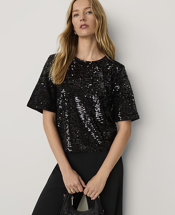 A woman wearing a black sequined top and black trousers stands against a plain white background.