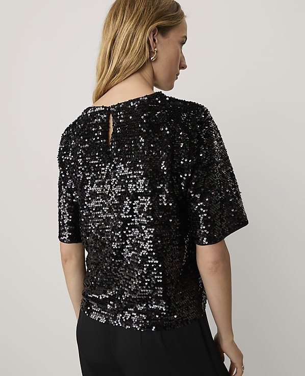 A woman wearing a black sequined top and black trousers stands against a plain white background.