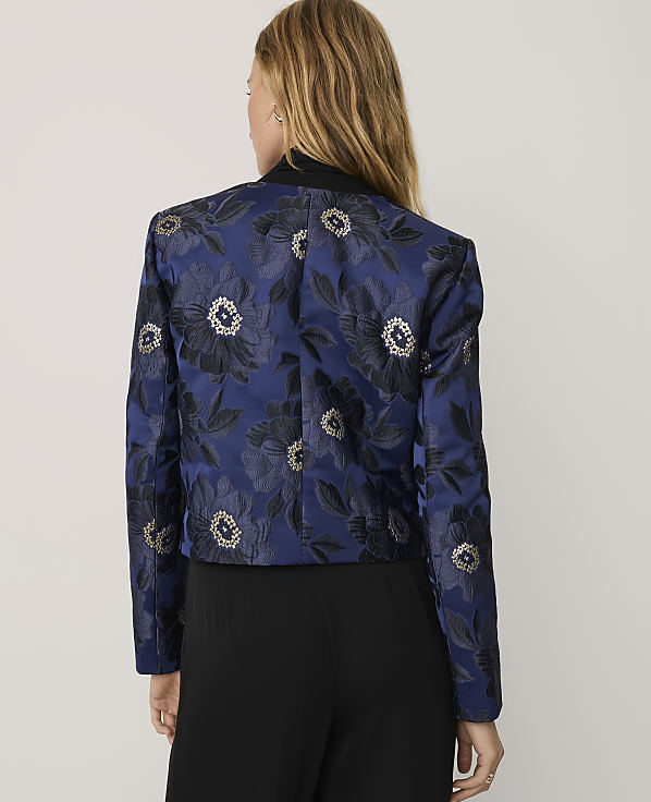A woman stands with her back to the camera wearing a blue floral jacquard blazer and black trousers against a plain light gray background.