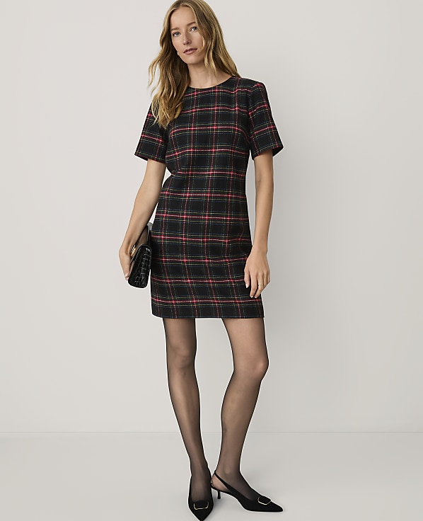A woman wearing a red and black plaid dress, black tights, and black heels stands against a plain white background.