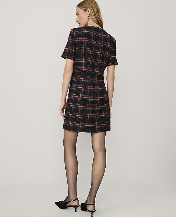 A woman wearing a red and black plaid dress, black tights, and black heels stands against a plain white background.