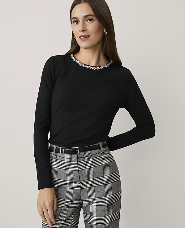 A woman wearing a black long-sleeved top with an embellished neckline and plaid trousers stands against a plain light grey background.
