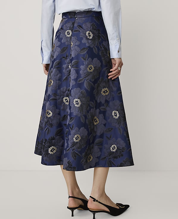 A person wears a light blue shirt and a navy blue floral jacquard skirt with black heels against a plain white background.