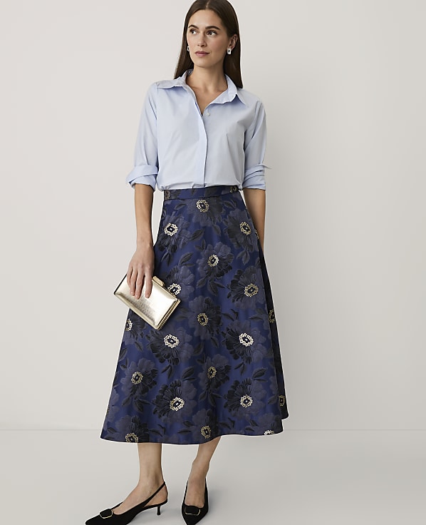 A woman wearing a light blue button-down shirt and a navy floral midi skirt stands against a plain white background.
