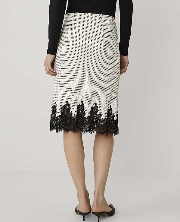A person stands against a plain white background wearing a black long-sleeved top and a white patterned skirt with black lace trim.