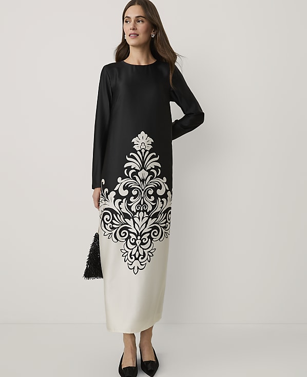 A woman wearing a black and white patterned midi dress and black heels stands against a plain white background.