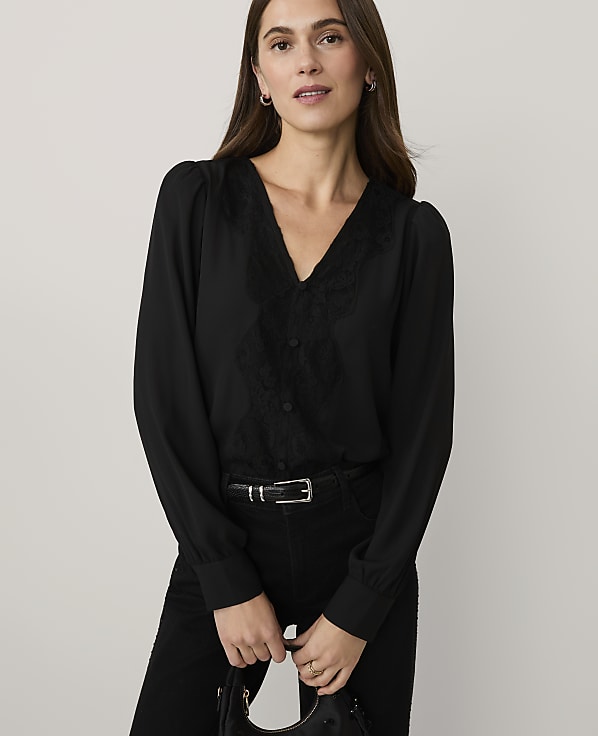 A woman wearing a black lace-detailed blouse and black trousers stands against a plain, light-colored background.