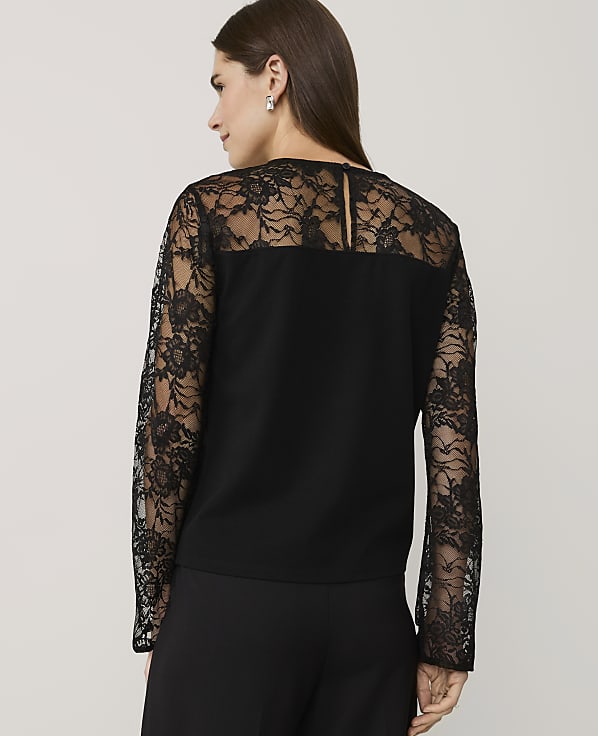 A woman wearing a black lace-detailed top and trousers stands against a plain, light-colored background.