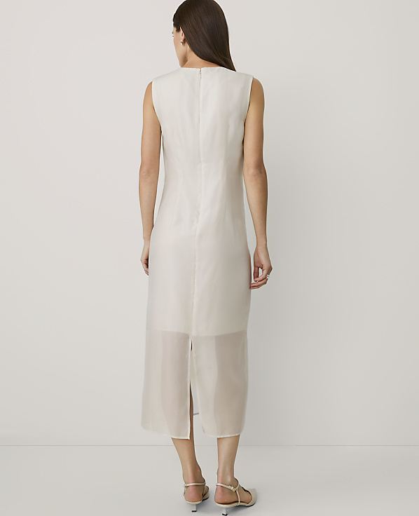 A woman stands with her back to the camera wearing a sleeveless white midi dress against a plain white background.