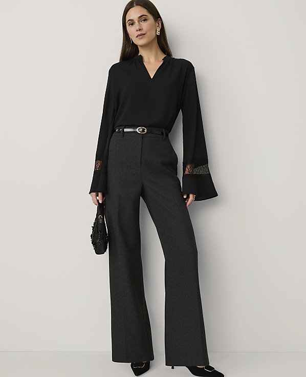 A woman wearing a black blouse and wide-leg grey trousers stands against a plain white background.