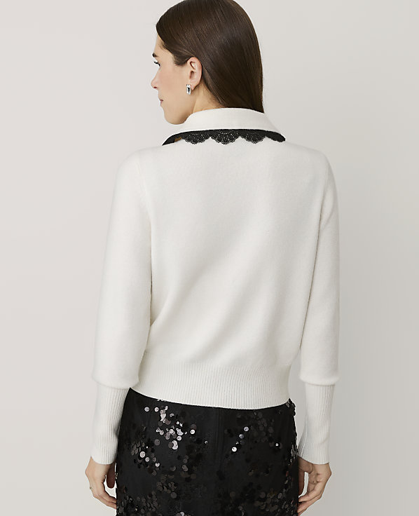 A woman wearing a white sweater with a black lace collar and sequined black pants stands against a plain light gray background.