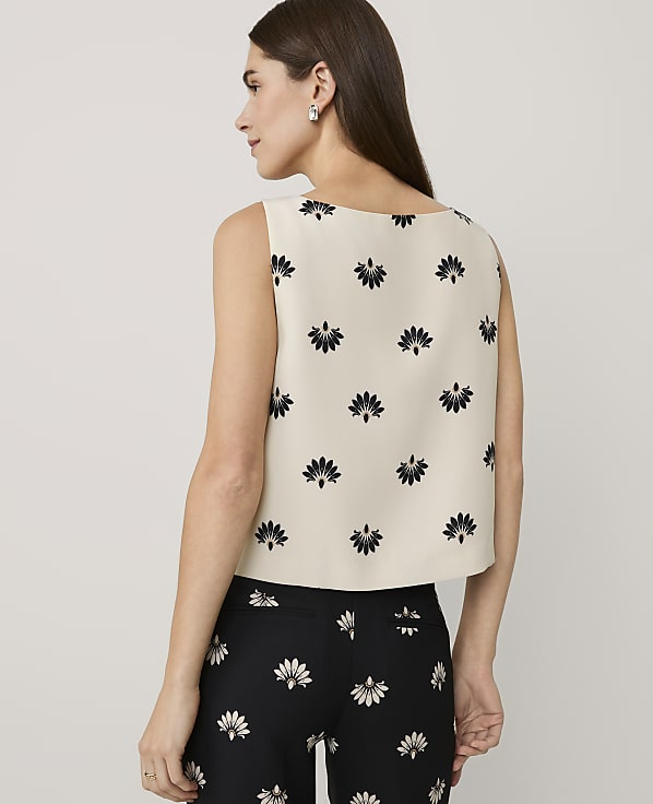 A woman stands with her back to the camera wearing a cream floral print top and black floral print pants against a plain light gray background.