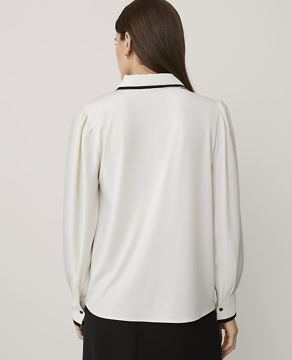 A woman wearing a white long-sleeved blouse and black trousers stands with her back to the camera against a plain light grey background.
