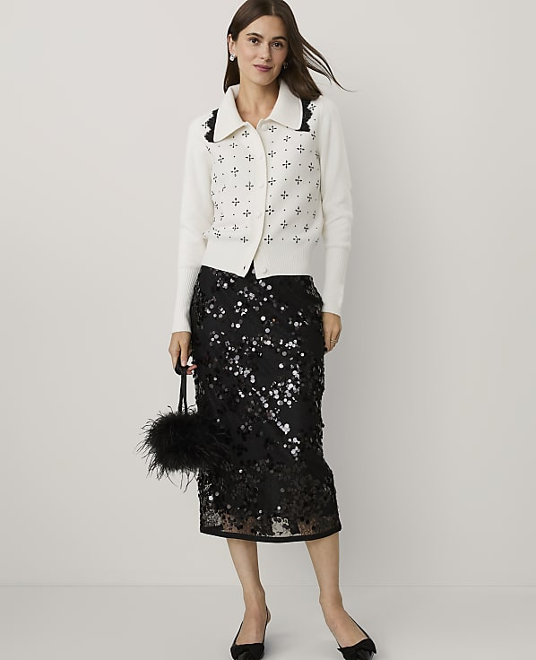 A woman wearing a white patterned cardigan and a black sequined skirt stands against a plain light gray background.