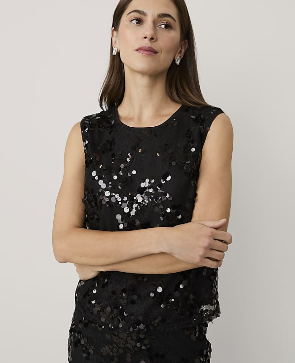 A woman wearing a black sequined sleeveless top stands with her arms crossed against a plain, light-colored background.