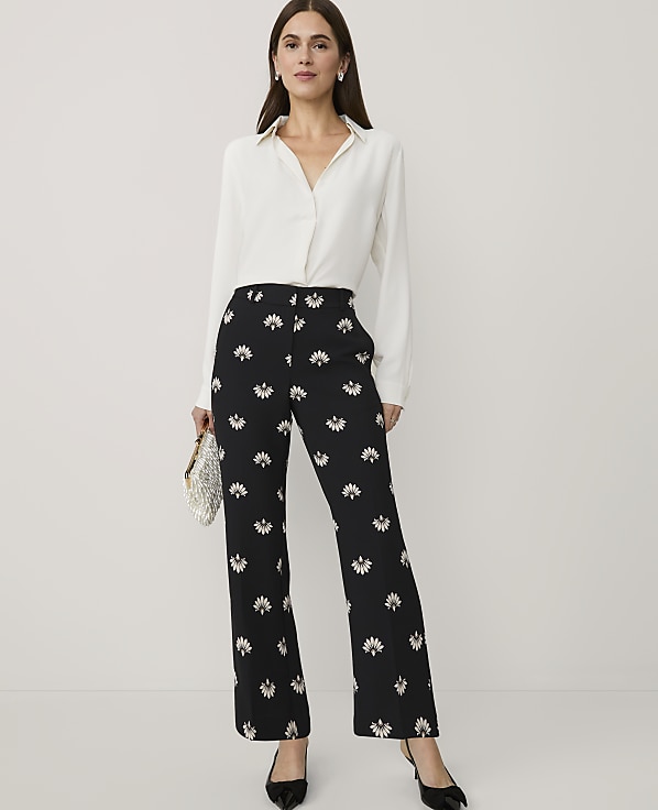 A woman wearing a white blouse and black floral trousers stands against a plain light grey background.