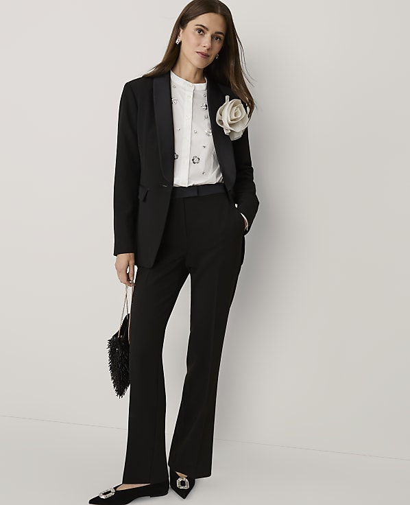 A woman wearing a black suit with a white embellished shirt and a large floral brooch stands against a plain white background.