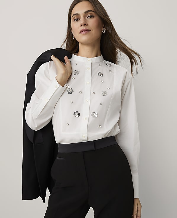 A woman wearing a white embellished shirt and black trousers stands against a plain light grey background, holding a black blazer over her shoulder.