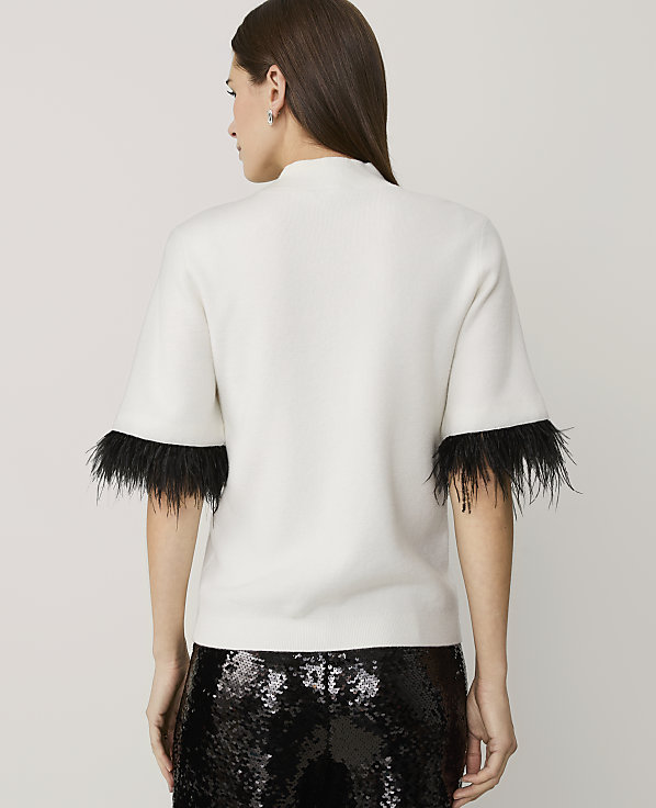 A woman stands with her back to the camera wearing a white short-sleeved top with black feather trim and sequined black pants against a plain light grey background.