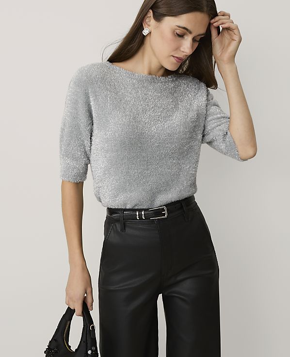 A woman wearing a shimmering silver sweater and black leather trousers stands against a plain light gray background.