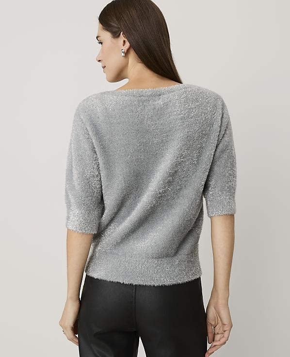 A woman wearing a grey fuzzy sweater and black leather pants stands with her back to the camera against a plain light grey background.