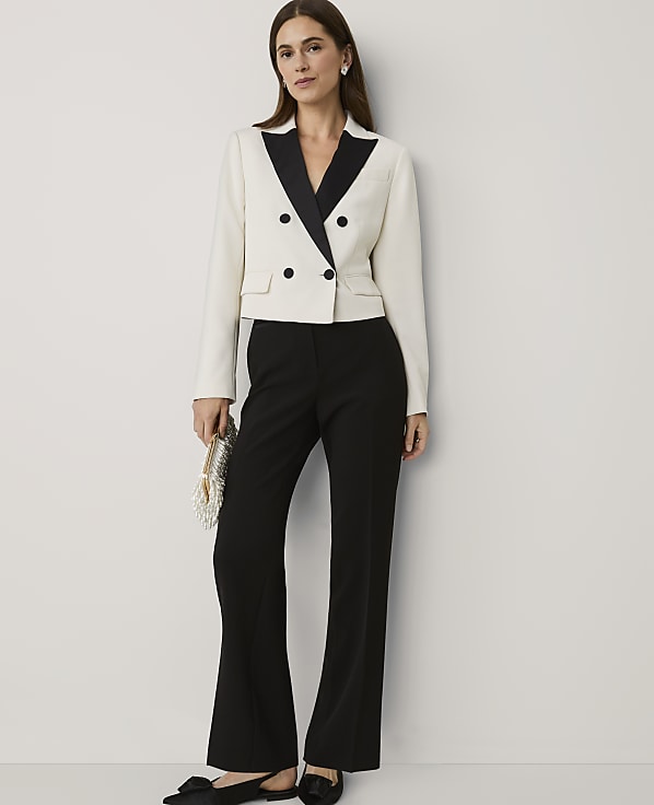A woman wearing a white and black tuxedo jacket with black flared trousers stands against a plain white wall.