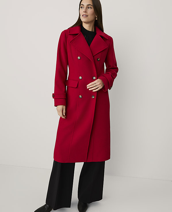 A woman wearing a long red double-breasted coat and black wide-leg trousers stands against a plain white background.