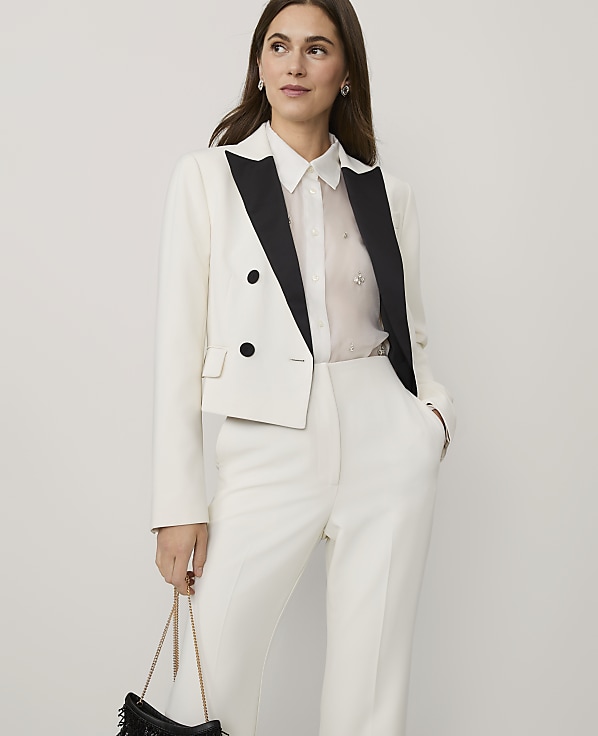 A woman wearing a white cropped blazer and matching trousers stands against a plain, light-colored background.