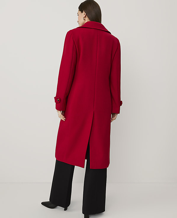 A woman wearing a long red coat and wide-leg black trousers stands with her back to the camera against a plain white background.