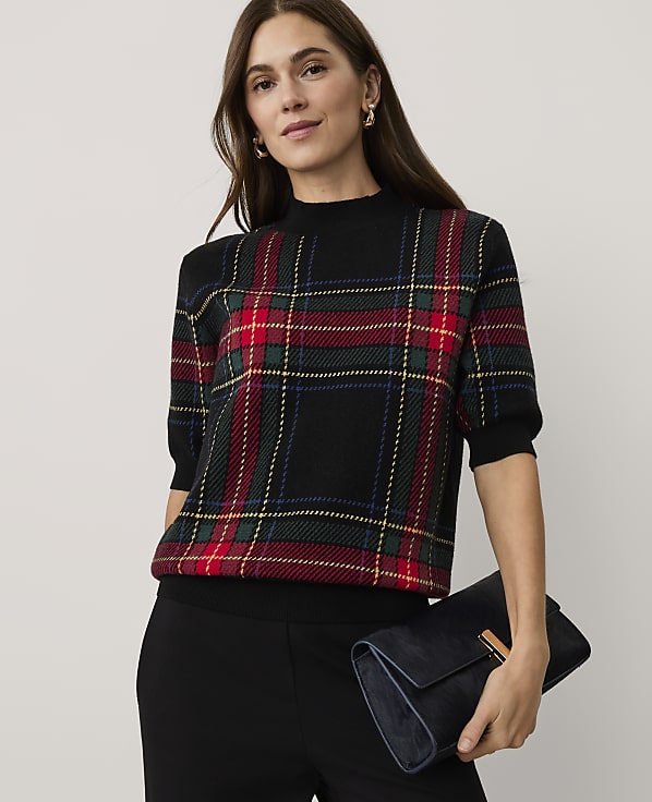 A woman wearing a red and green plaid sweater and black pants holds a dark blue clutch against a plain light grey background.