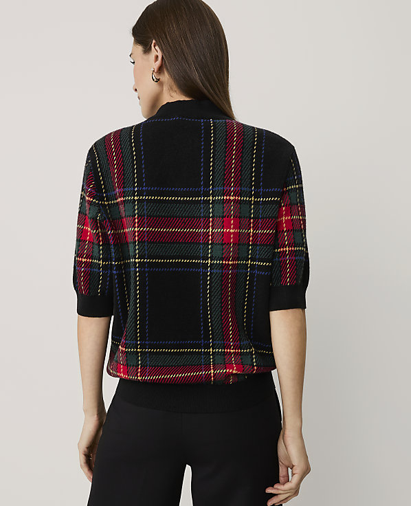 A woman stands with her back to the camera wearing a plaid short-sleeved sweater and black trousers against a plain light gray background.