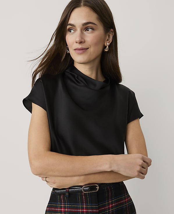 A woman wearing a black satin top and plaid trousers stands against a plain, light-colored background.