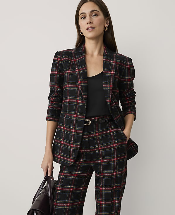 A woman wearing a red and black plaid suit stands against a plain, light-colored background.